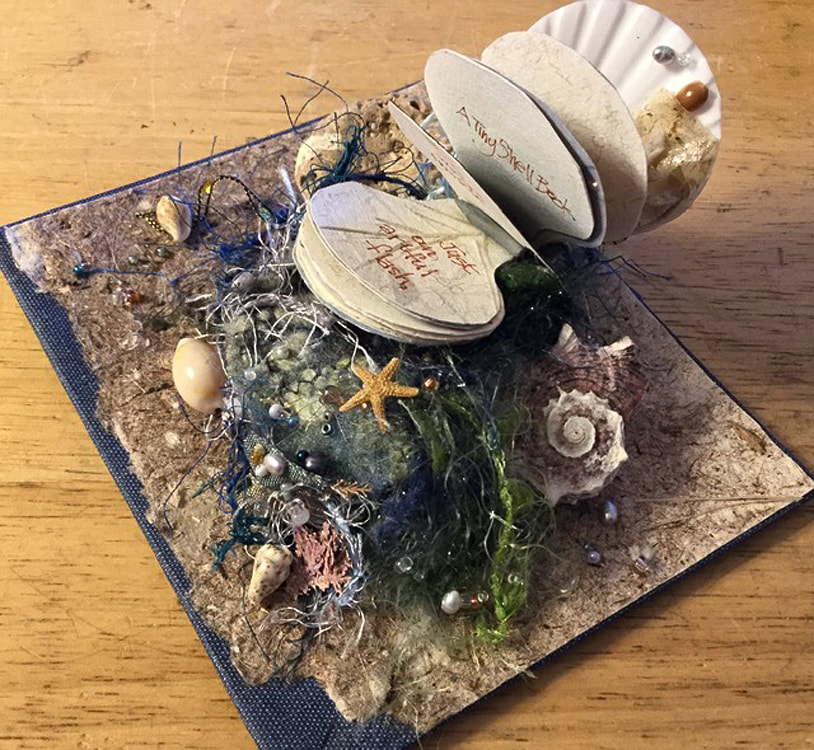 A miniature artist’s beach book. The shell itself is about 1-1/2” wide, book contains one of my poems in calligraphy, multiple art papers, lots of tiny engineering. The base is a covered book board, handmade paper and a needle felted support with yarn, threads, shells, and a rock.
